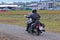 A motorcyclist in leather outfit rides a chopper on a dirt road along a dirt road against a backdrop of urban infrastructure. Hobb