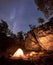 Mountain campsite at night amid huge steep rock formation. Tourist tent lit by burning campfire