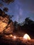 Mountain campsite at night amid huge steep rock formation. Tourist tent lit by burning campfire