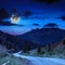 Mountain road near the coniferous forest with cloudy moon sky