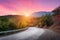 Mountain road passing through the forest with dramatic colorful sky and red clouds at colorful sunset in summer. Mountain