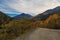 A mountain road on a sharp turn with a view of the autumn multicolored forest, mountain peaks and feathery clouds against the blue