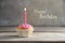 Muffin or cupcake with a pink burning candle on a wooden board against a rustic background, text Happy Birthday, greeting card