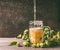 Mug of beer on a rustic table with a vine and cones of hops opposite a dark wall, front view.