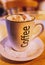 Mug of cappuccino coffee with the word coffee written on the side. On a saucer with a paper lace doily on a round wooden table