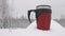 Mug thermos standing in a snowy forest in the heavy snowfall. Dark red in color with black handle