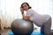 Multi ethnic adult pregnant woman leaning on a fit ball, smiles at camera, doing prenatal fitness exercises at home.