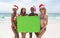 Multi-ethnic female friends holding a empty green placard at beach on a sunny day