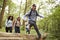 Multi ethnic group of five young adult friends running in a forest and jumping over fallen tree during a hike