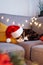 Multicolored relaxed cat lying on a gray sofa in a Santa& x27;s hat with blurred Christmas decor composition on the