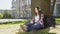 Multinational girl sitting under tree in headphones, using phone, choosing song