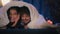 Multiracial couple man and woman laughing smiling happy cover heads with blanket under duvet at night evening home using