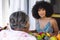 Multiracial smiling young woman with afro hair talking with senior woman at dining table at home