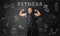 Muscled young man showing his bicep muscles on the background of blackboard with fitness doodles