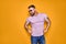 Muscular confident bearded male in shirt, jeans and stylish sunglasses standing in studio over yellow background