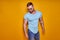 Muscular confident bearded male in shirt, jeans and stylish sunglasses standing in studio over yellow background