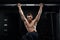 Muscular male athlete pulling up on horizontal bar in dark gym.