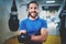 Muscular smiling boxer in gloves standing with arms crossed near punching bag in fitness gym on blurred background.Young