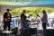 Musical group is seen from the back as they play their instruments in front of a crowd at an outdoor fair. There is a woman