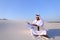 Muslim male architect sitting with laptop on sand in desert on h