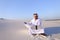 Muslim male architect sitting with laptop on sand in desert on h