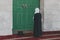 Muslim religious woman in long dress and covered head standing against the massive green door of mosque and praying