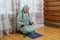 A Muslim woman in a light dress for prayer prays while sitting on a mat