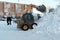 A Mustang skid steer pours snow into a pile outside after a snowfall with residential buildings in the background