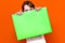 Mysterious caucasian young girl hiding under bright green folder with a banner advertisement on a orange
