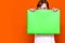 Mysterious caucasian young girl hiding under bright green folder with a banner advertisement on a orange