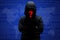 Mysterious unknown hacker in black clothing with hoodie, stands crossed hands against dark digital interface background with red q