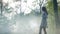 Mysterious woman wearing white dress walks in the mist fog in the woods at sunrise -