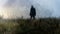 A mysteryious figure wearing a long coat and fedora hat, with back to camera, standing in a field. With an artistic, blurred,