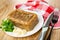 Napkin, piece of meat jelly, basil, garlic in white plate, knife and fork on wooden table