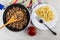 Napkin, spoon in frying pan with fried mince, white plate with pasta, fork, bowl with ketchup on table. Top view