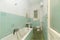 Narrow bathroom with green tiles and dirty floors, bathtub and white toilets