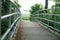 A narrow bridge with metal railings on both sides surrounded by lush green vegetation diminishing in the perspective