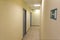 A narrow light corridor in the office room with several gray doors and signs.