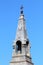 Narrow light grey metal gothic church tower with various decorative elements and rusted cross on top