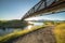 Narrow road along a shimmering lake with bridge and blue sky overhead