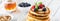 Narrow view of a stack of buttermilk pancakes topped with berries and banana slices and served with syrup and walnuts.