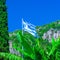 National flag of Greece on the flagpole against of bananas leaf and cypress tree and green mountain slopes. Mediterranean sea,