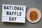 `National Waffle Day` word on lightbox, traditional belgian waffle on pink plate over concrete background,