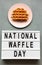 `National Waffle Day` word on lightbox, traditional belgian waffle on pink plate over concrete background, top view. From above