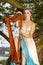 Natural Portrait of Cute Caucasian Blond Girl in Long Dress Playing the Harp on tree Branch on Park Outdoors