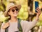 Nature, phone selfie and woman hiking with hat, sunglasses and smile, adventure in forest or woods in summer. Freedom