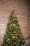 Near the brick wall is a beautiful new year`s green pine tree, decorated with different balls, flowers and a garland on the wall