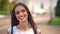 Neautiful woman is standing on bus stop in daytime in summer, watching at camera, smiling, blurred road on background