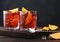 Negroni cocktail in crystal glasses with orange slice and fresh raw oranges on chopping board with spoon on dark wooden background