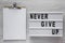 `Never give up` words on lightbox, clipboard with blank sheet of paper over white wooden background. Flat lay, from above,